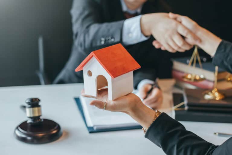 Two people are shaking hands at a desk. One person holds a small model house, suggesting a property deal in progress, while legal items such as a gavel, scales of justice, and documents scatter the surface. The presence of a construction lawyer could be inferred in this mid-level negotiation setting.