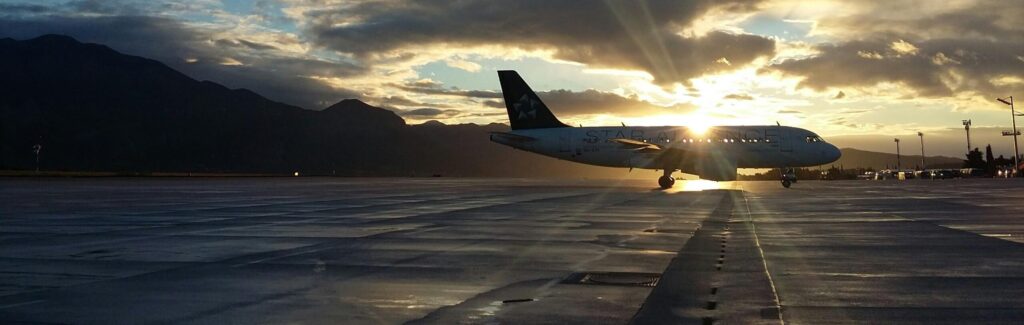 An airplane is parked on a tarmac at sunset in Australia, with the sun partially hidden behind the aiRostron Carlyleaft and mountains visible in the background.